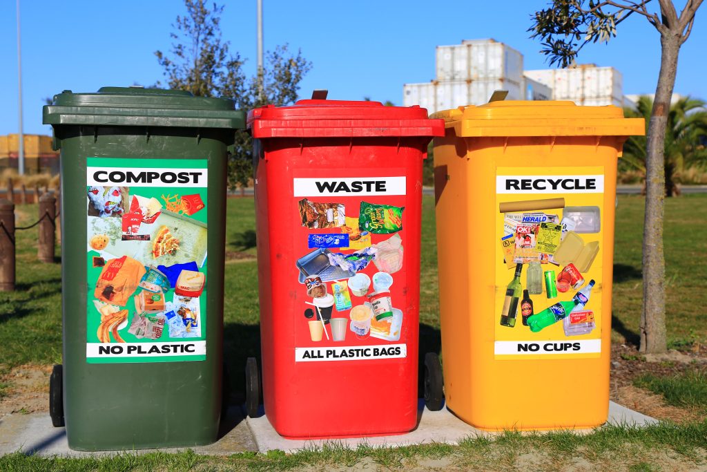 Three colorful bins, labelled "Compost," "Waste," and "Recycle" sit side by side. Each bin's label shows photos of materials that should be placed inside.